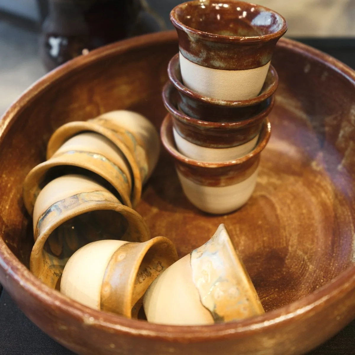 A brown glazed bowl holding small handmade cups, some stacked, some resting on their sides.