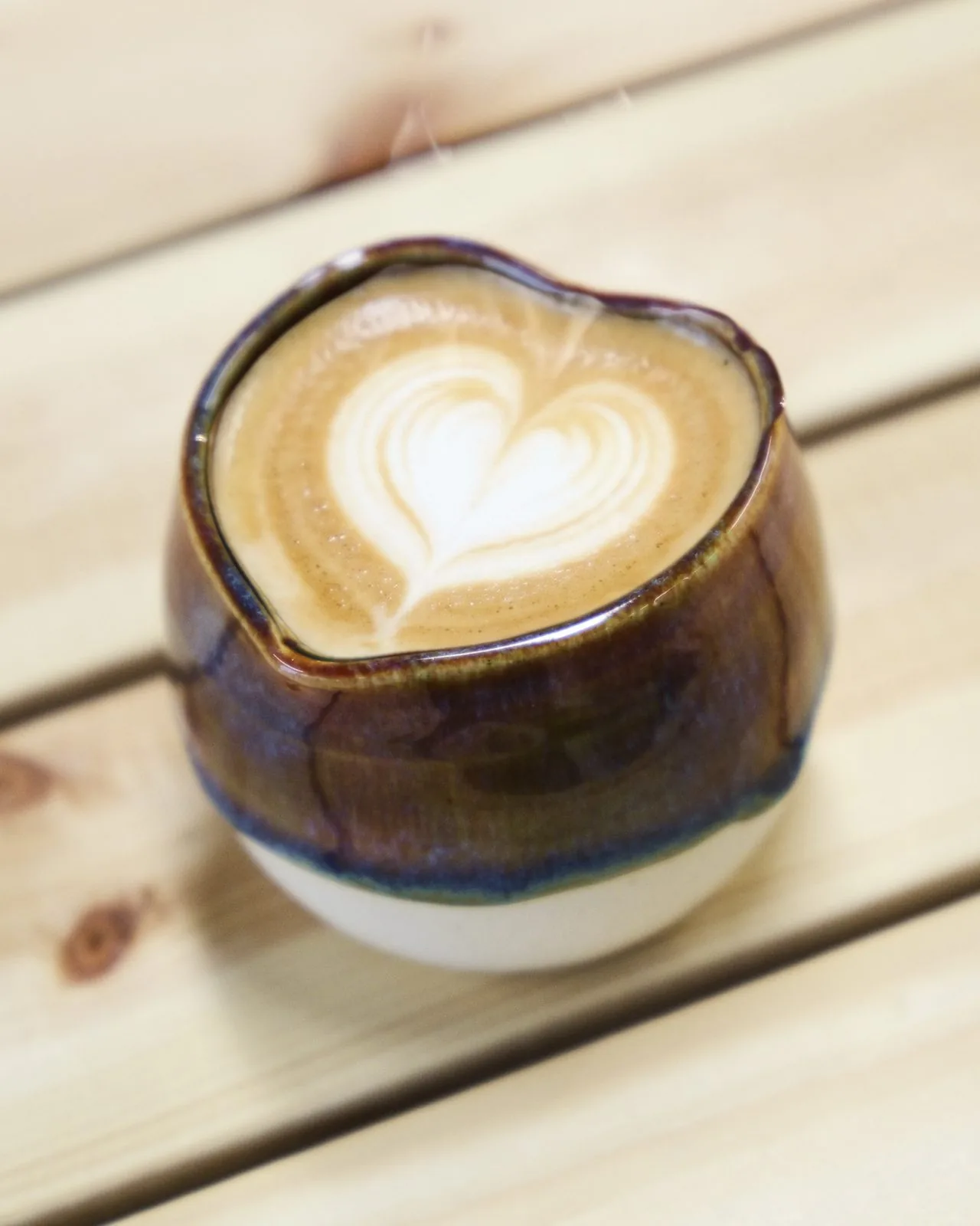 A latte with heart latte art in a handmade heart-shaped cup with a glossy brown and amber glaze.