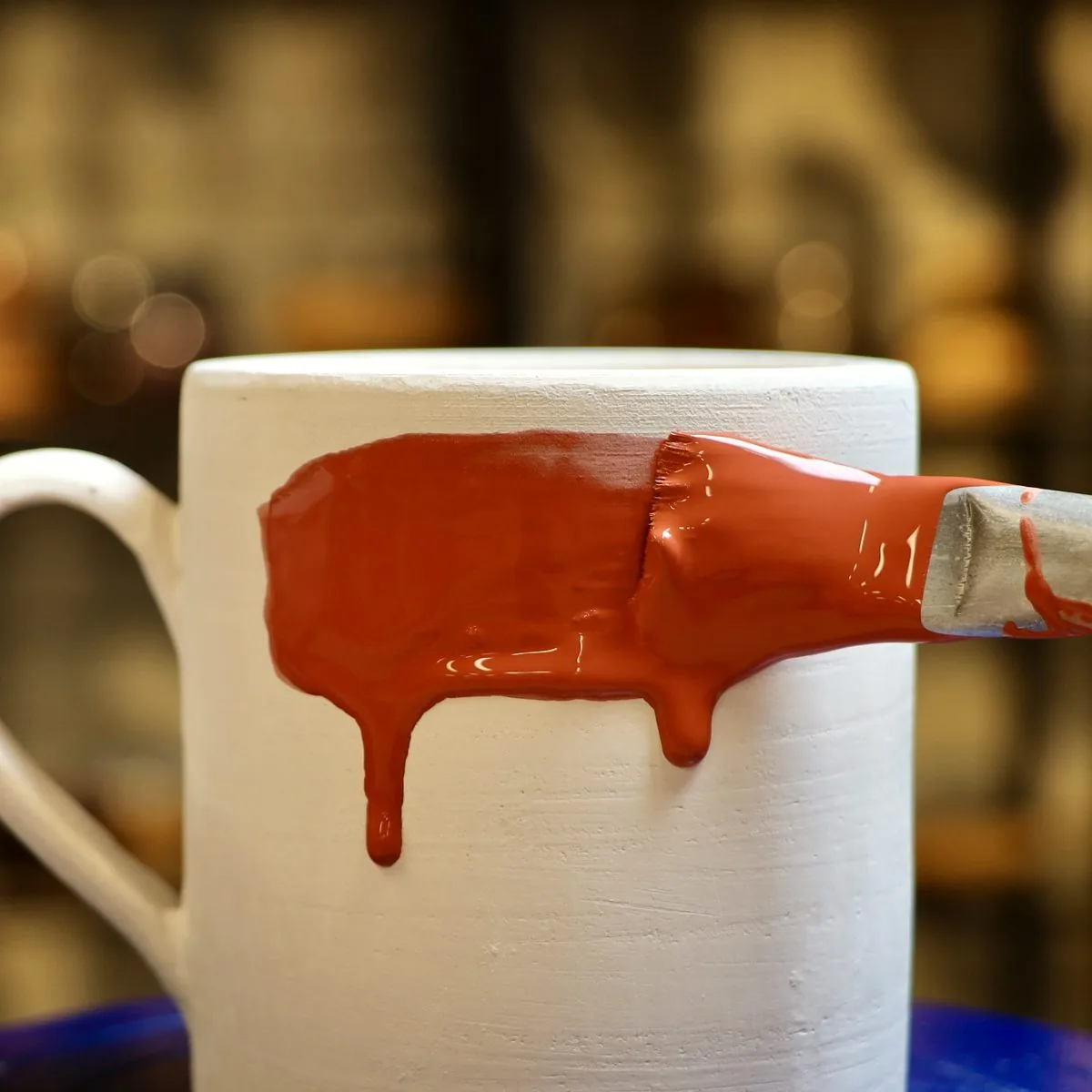 Glazing: a brush laying terracotta-red glaze on a white bisque mug, the glaze running in drips.
