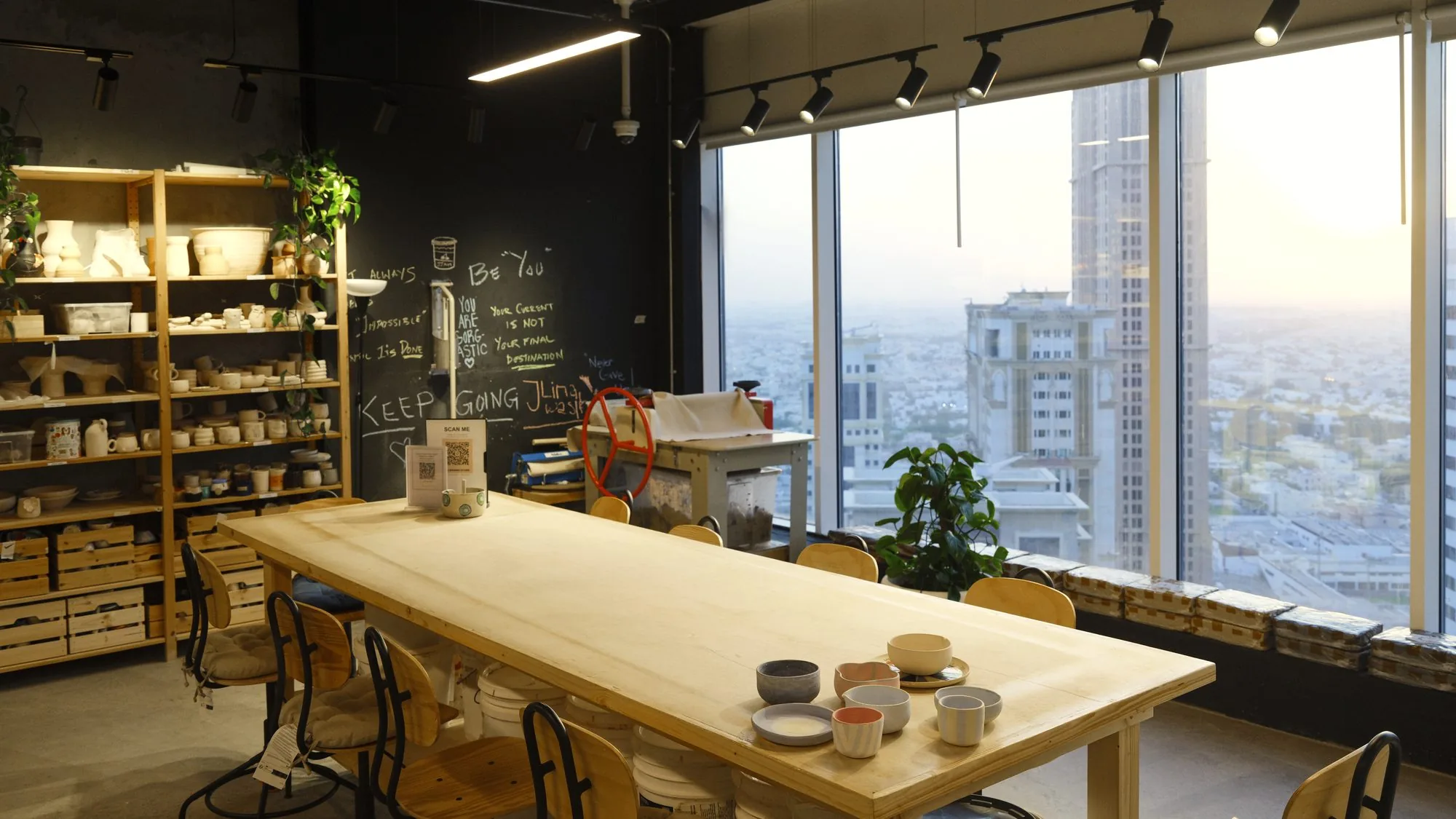 The studio: a long wooden worktable with handmade cups, shelves of pottery and tall windows over the city at sunset.